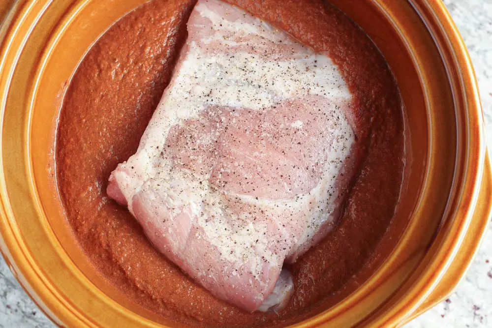 raw pork shoulder seasoned with salt and pepper and added to a brown slow cooker pot filled with red mole sauce
