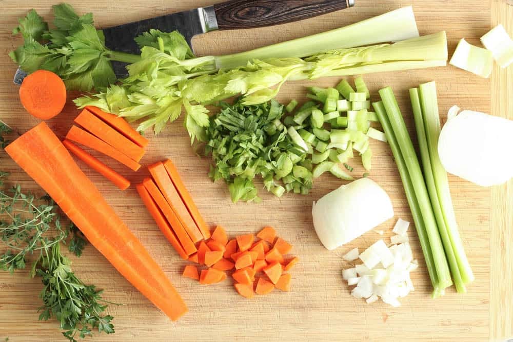 green celery, orange carrots, white onion with cilantro and thyme being diced and prepared fo recipe on a light wooden cutting board
