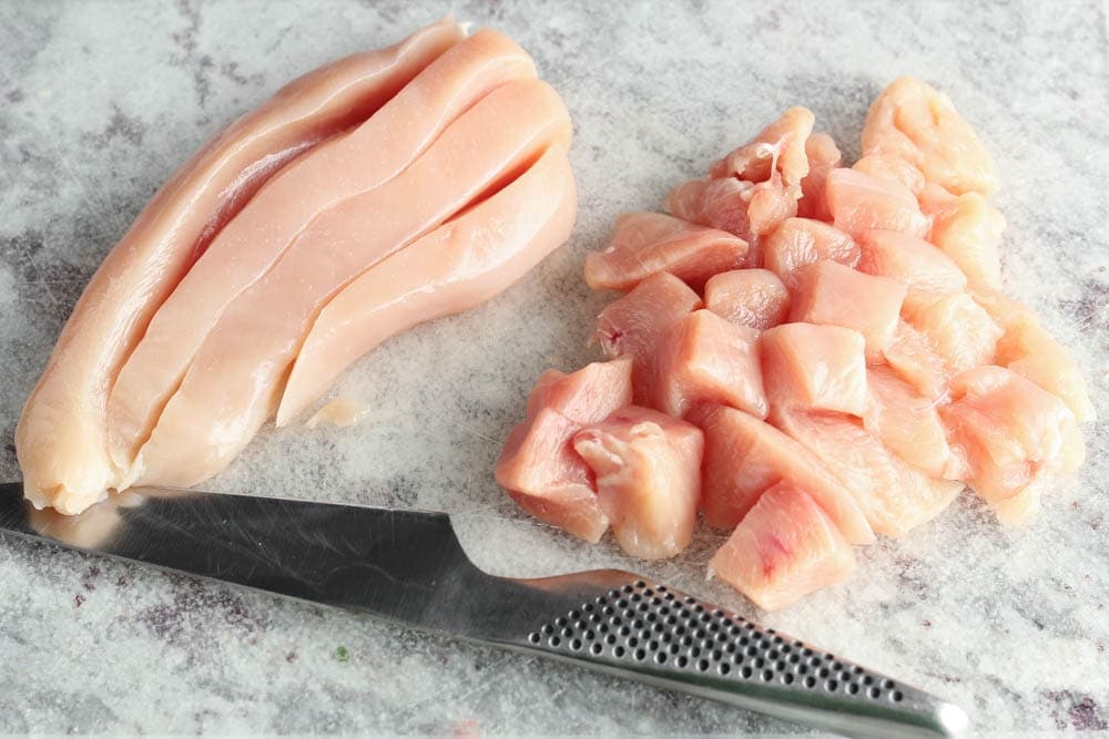 raw chicken breast on a plastic clear cutting board being cut into one inch chunks