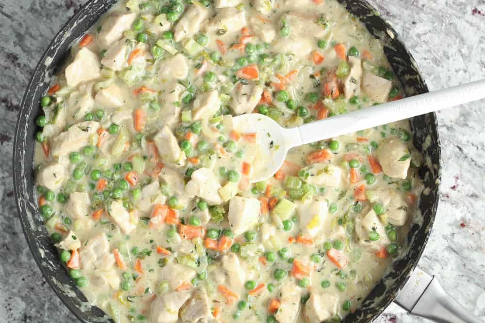 large black saute pan filled with creamy chicken filling consisting of diced chicken, carrots, peas, celery and onion being stirred with a white spatula