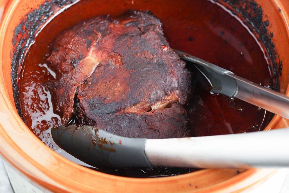 Pork shoulder in a slow cooker after cooking for 24 hours in a deep red mole sauce with tongs reaching in to grab it