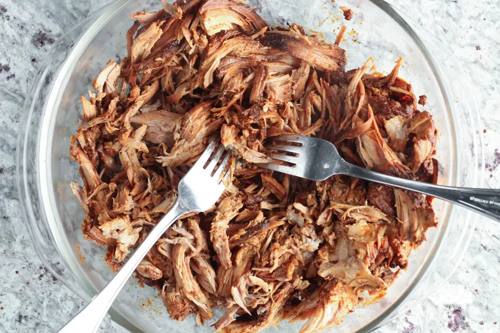 Cooked pork sholder being shredded with two silver forks in a clear glass bowl