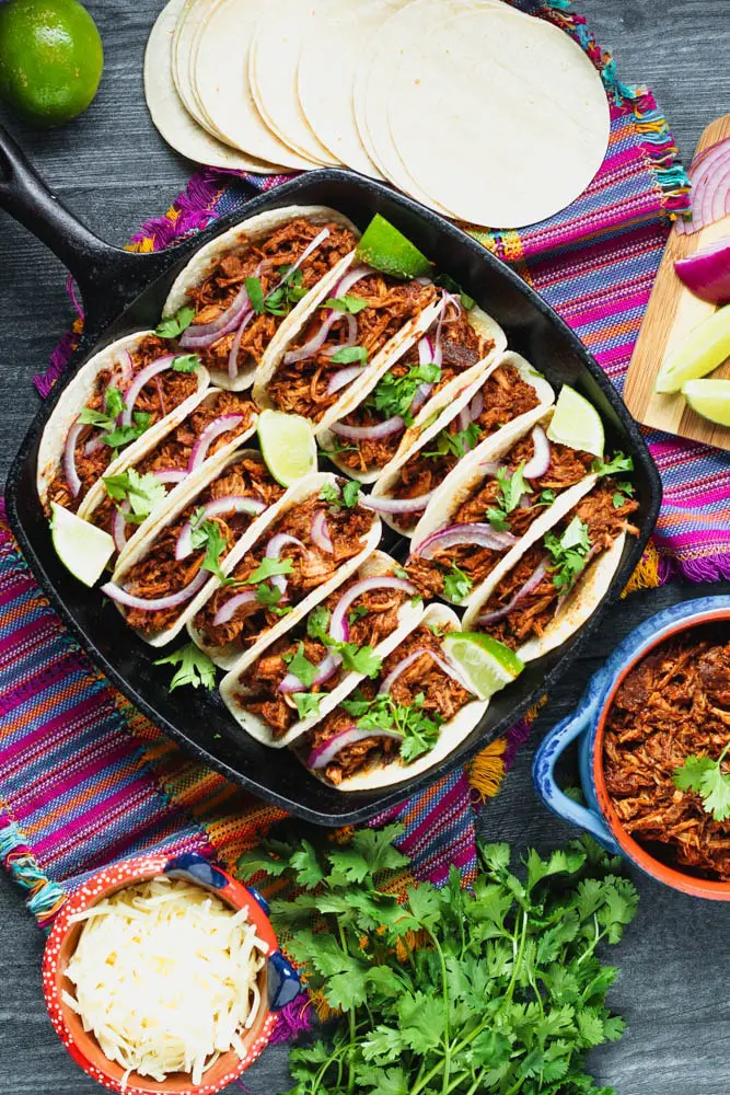 a dozen mole pulled pork tacos in a cast iron pan topped with red onions and cilantro surrounded by a bright purple napkin, cilantro leaves, corn tortillas and extra shredded pork in a blue bowl