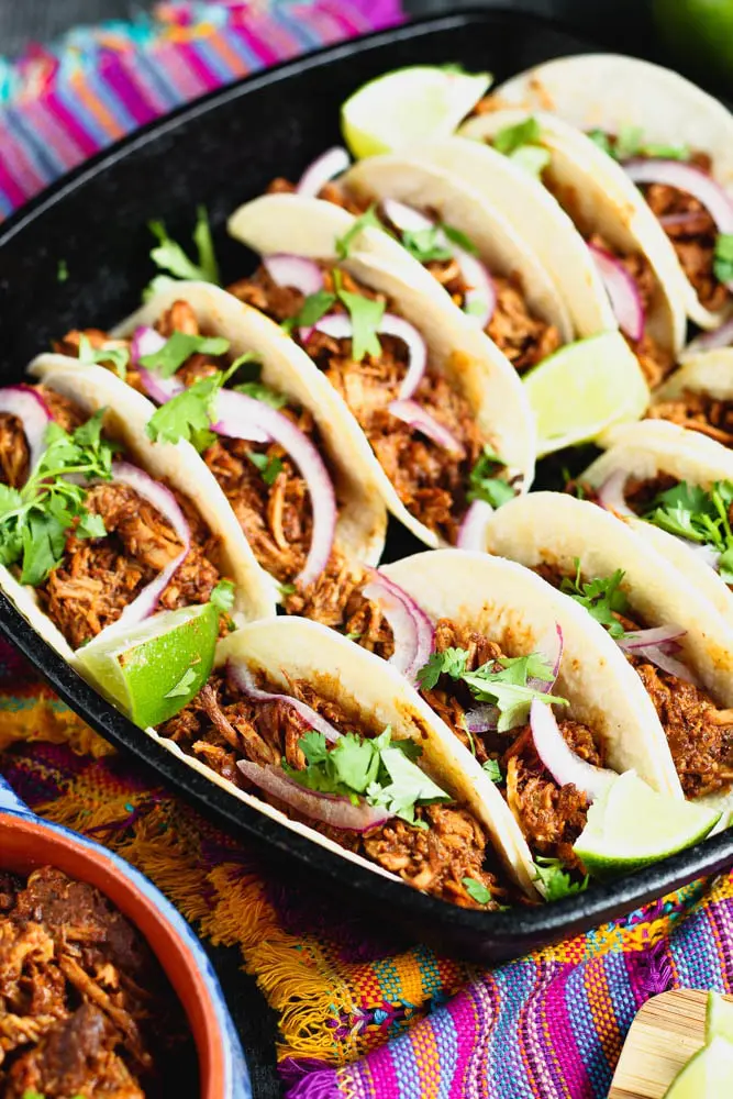mole pork tacos topped with thinly sliced red onion, bright green cilantro and lime wedges on a dark black plate