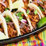 5 mole pork tacos topped with thinly sliced red onion, bright green cilantro and lime wedges on a dark black plate