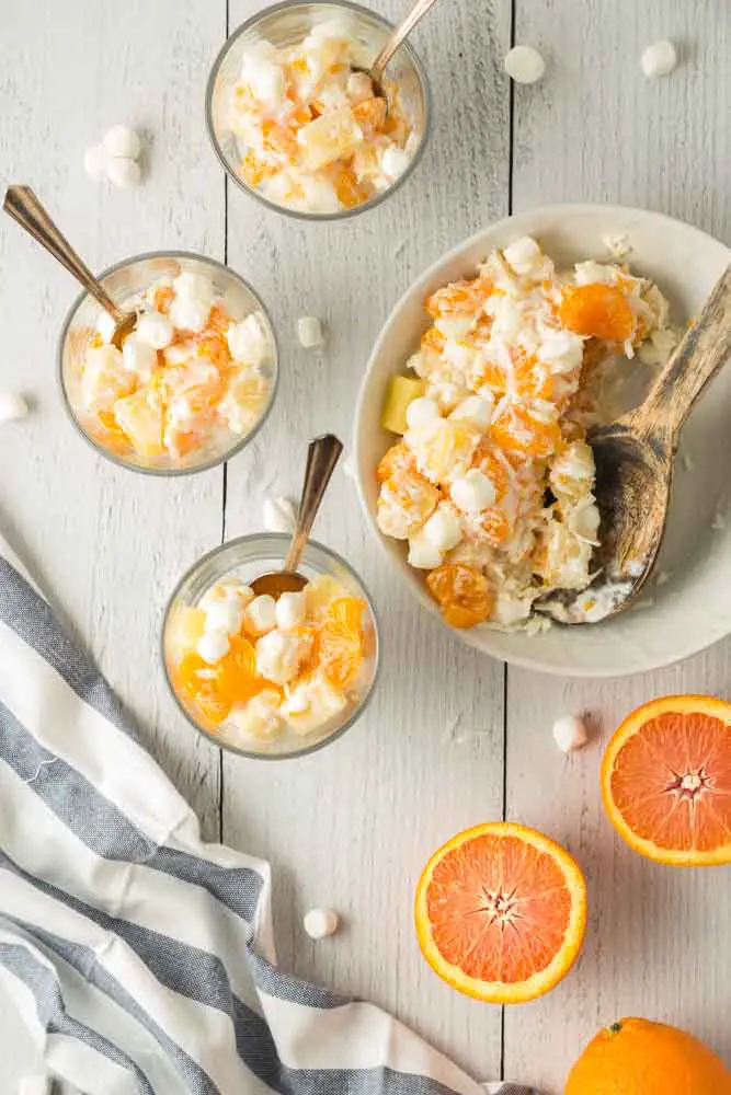 Five Cup Salad being scooped from a large white bowl with a wooden spoon and served into three small clear glass trifle dishes and two orange halves