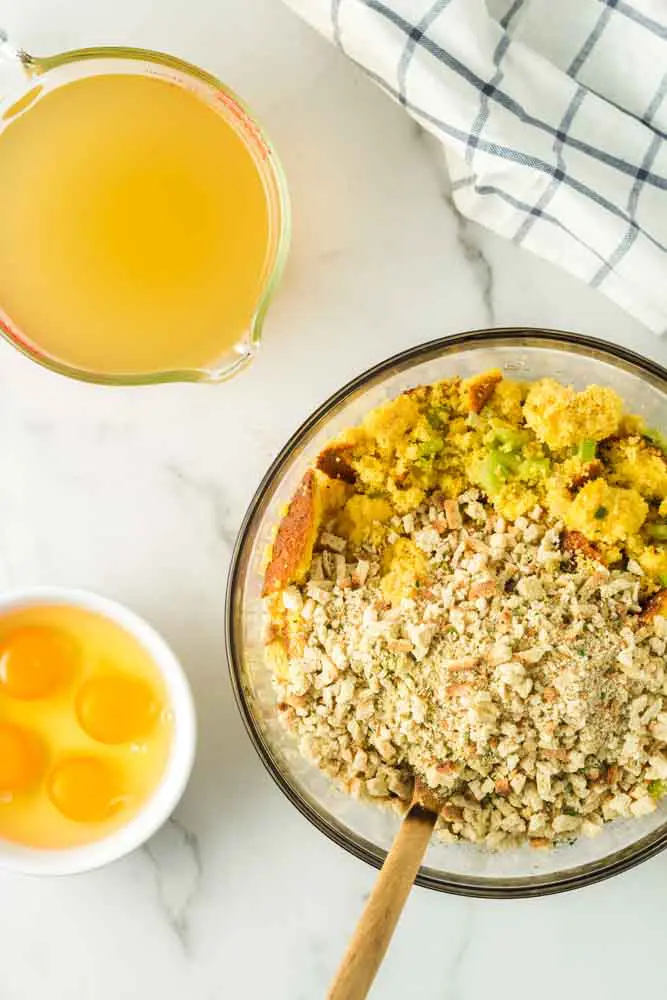 cornbread and vegetable mixture in a glass mixing bowl with a wooden spoon and the chicken stock and eggs on the side