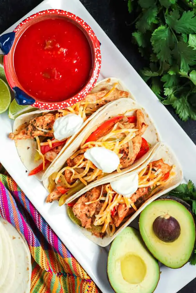 Three prepared chicken fajitas tacos with slices of chicken, peppers, onions, shredded cheese and a dollop of sour cream with a bowl of red salsa and fresh green avocadoes on the side