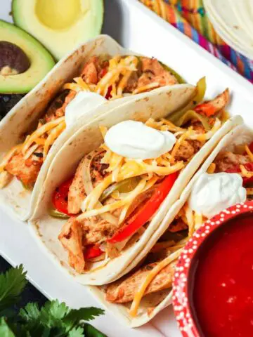Three prepared chicken fajitas tacos with slices of chicken, peppers, onions, shredded cheese and a dollop of sour cream with a bowl of red salsa and fresh green avocadoes on the side