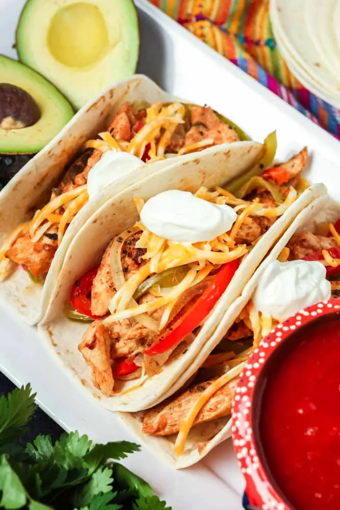 Three prepared chicken fajitas tacos with slices of chicken, peppers, onions, shredded cheese and a dollop of sour cream with a bowl of red salsa and fresh green avocadoes on the side