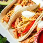 Three prepared chicken fajitas tacos with slices of chicken, peppers, onions, shredded cheese and a dollop of sour cream with a bowl of red salsa and fresh green avocadoes on the side