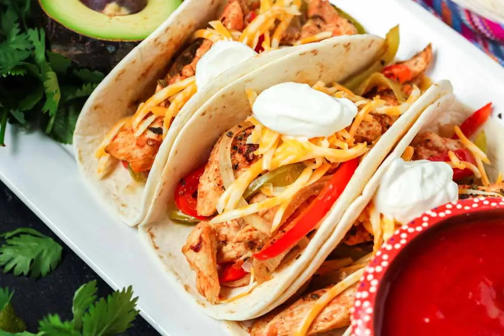 Three prepared chicken fajitas tacos with slices of chicken, peppers, onions, shredded cheese and a dollop of sour cream with a bowl of red salsa and fresh green avocadoes on the side