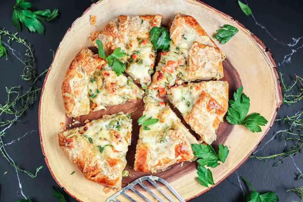 Cooked chicken pot pie crostata sliced into 6 triangular pieces sitting on a round wooden serving platter on a black backdrop with green parsley leaves scattered around