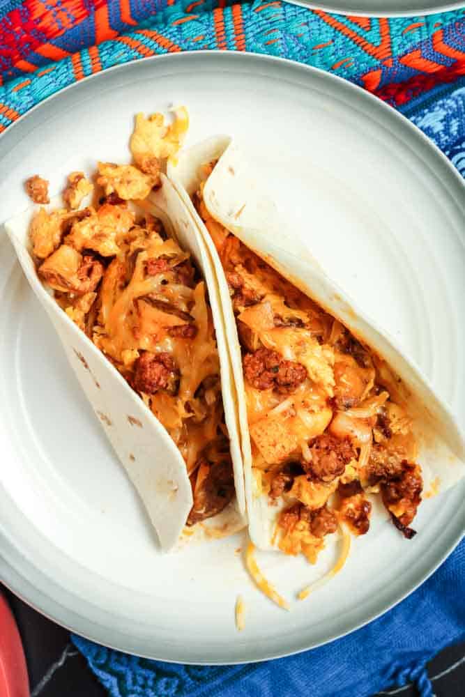Two chorizo and potato breakfast tacos filled with orange chorizo, yellow scrambled eggs and chunks of potato and onion served in a flour tortilla on a white plate and a festive Mexican blanket backdrop