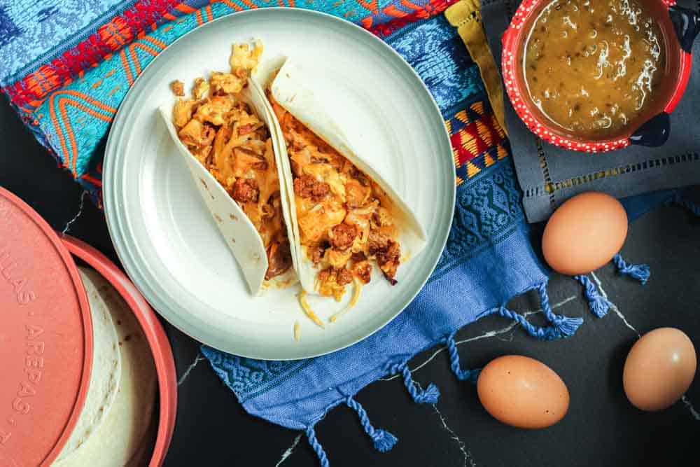 Two chorizo and potato breakfast tacos filled with orange chorizo, yellow scrambled eggs and chunks of potato and onion served in a flour tortilla on a white plate and a festive Mexican blanket backdrop