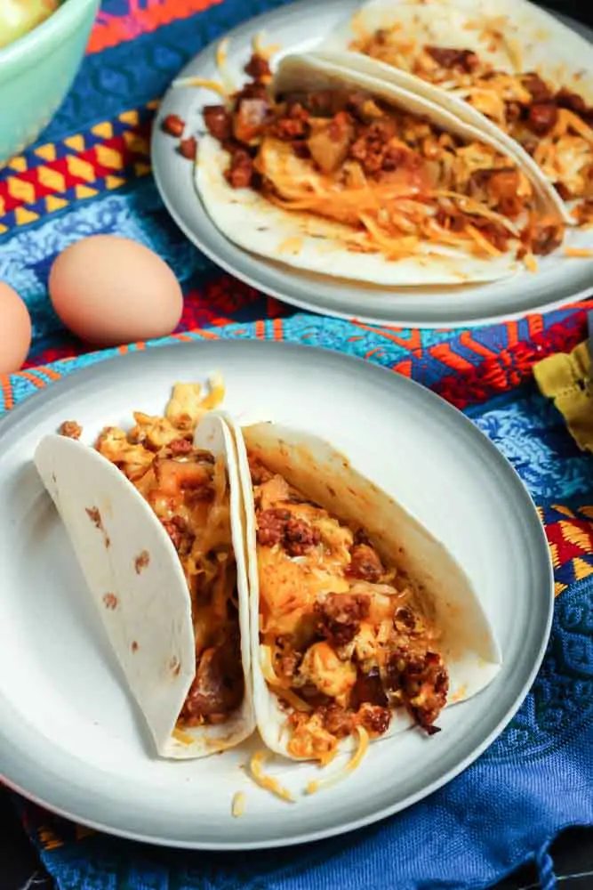 Two chorizo and potato breakfast tacos filled with orange chorizo, yellow scrambled eggs and chunks of potato and onion served in a flour tortilla on a white plate and a festive Mexican blanket backdrop