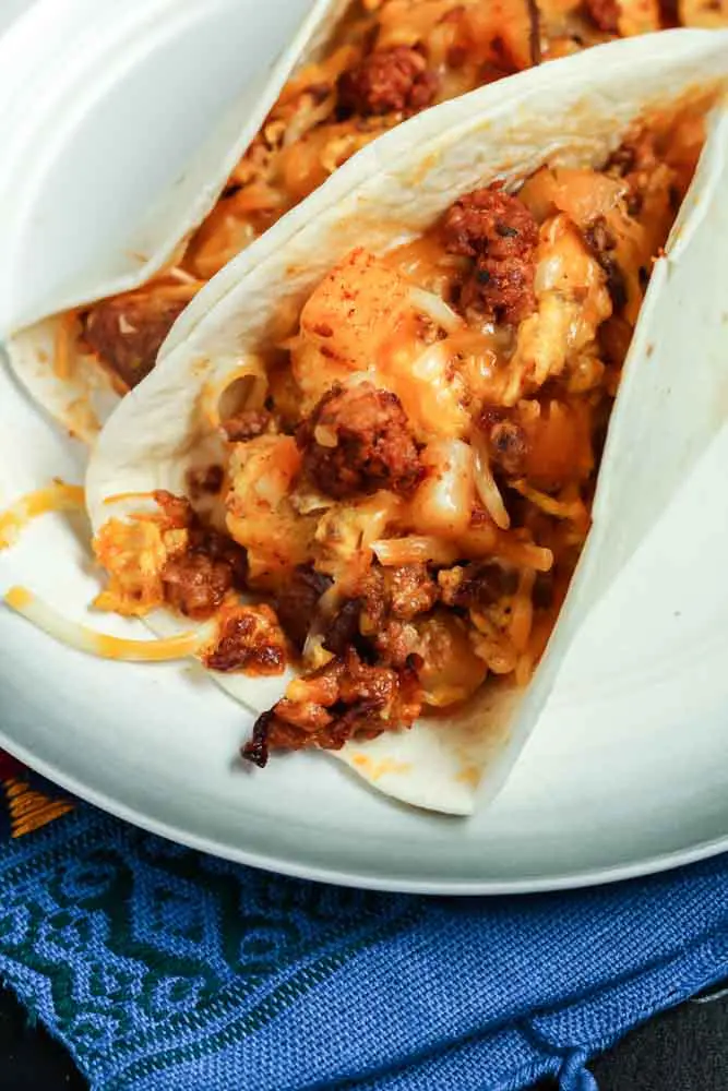 Two chorizo and potato breakfast tacos filled with orange chorizo, yellow scrambled eggs and chunks of potato and onion served in a flour tortilla on a white plate and a festive Mexican blanket backdrop