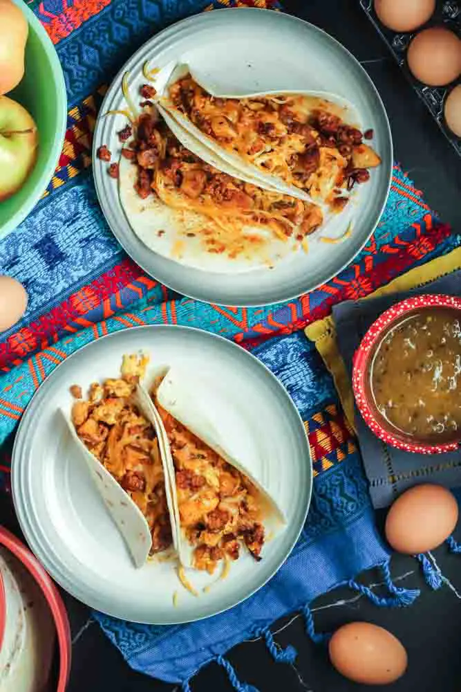 Two chorizo and potato breakfast tacos filled with orange chorizo, yellow scrambled eggs and chunks of potato and onion served in a flour tortilla on a white plate and a festive Mexican blanket backdrop