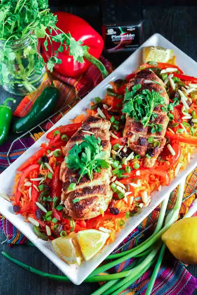 white rectangular plate filled with orange rice topped with strips of red bell pepper, green onion pieces, sliced chicken breast and cilantro with a lemon wedge