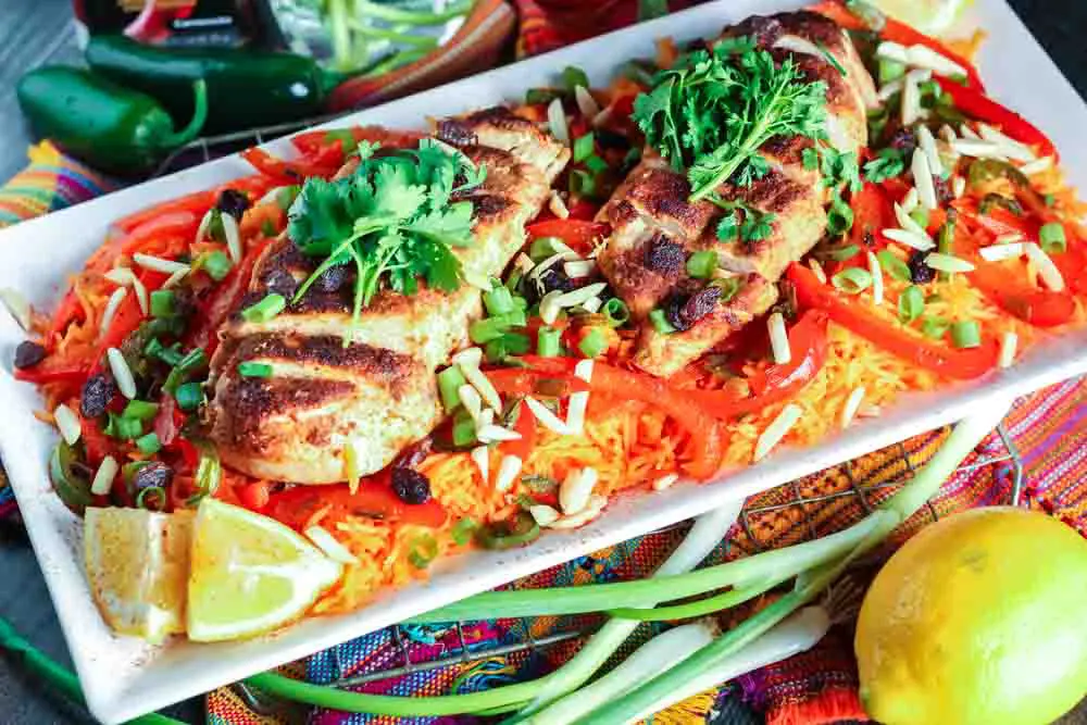 white rectangular plate filled with orange rice topped with strips of red bell pepper, green onion pieces, sliced chicken breast and cilantro with a lemon wedge