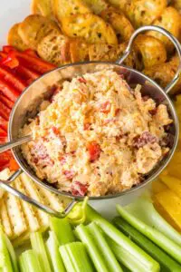 close up of silver bowl of orange pimento cheese surrounded by green celery sticks, red and yellow bell pepper strips and golden brown toasted baguette