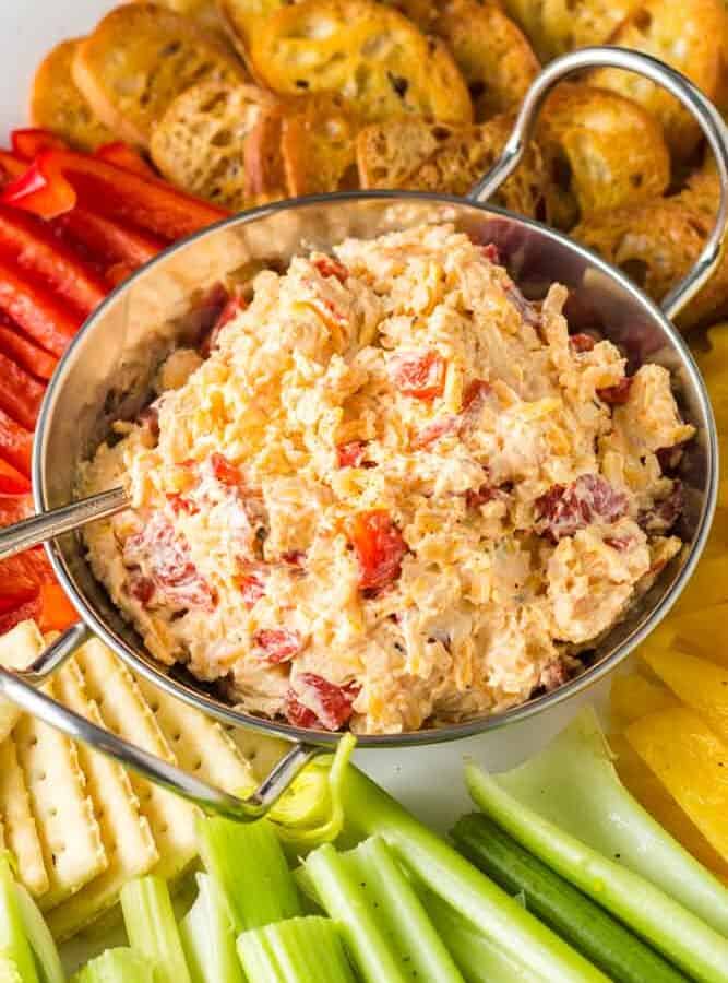 close up of silver bowl of orange pimento cheese surrounded by green celery sticks, red and yellow bell pepper strips and golden brown toasted baguette