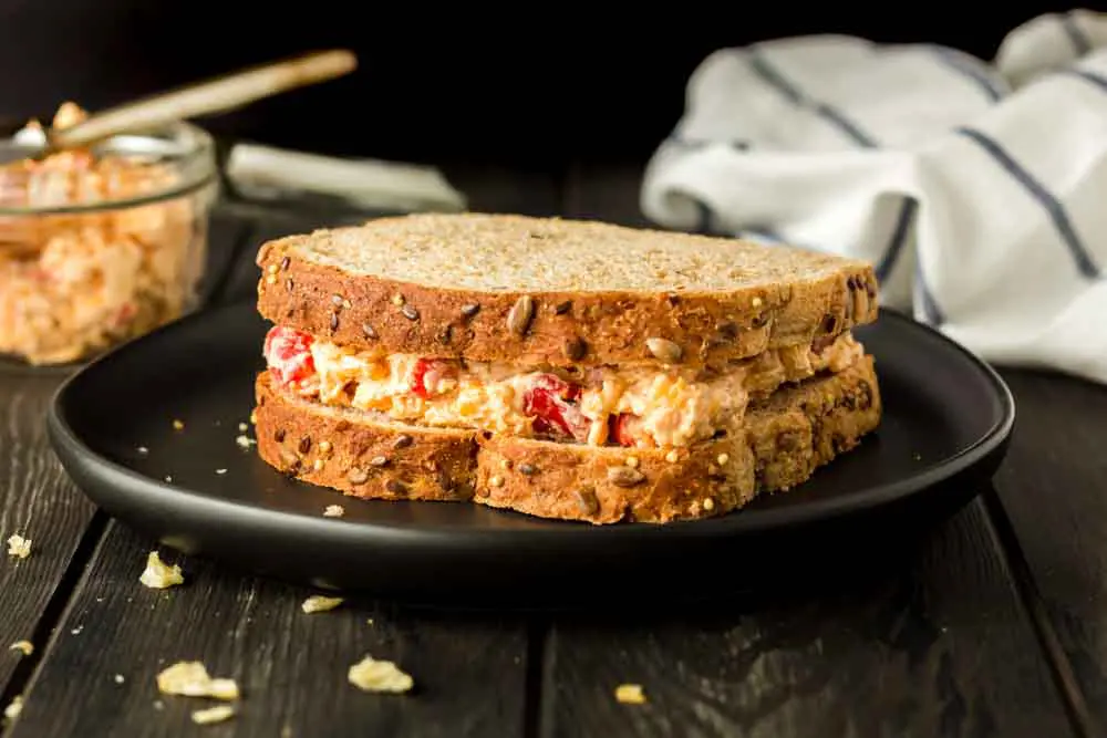 Pimento cheese sandwich on wheat bread on a black plate with bread crumbs on the board