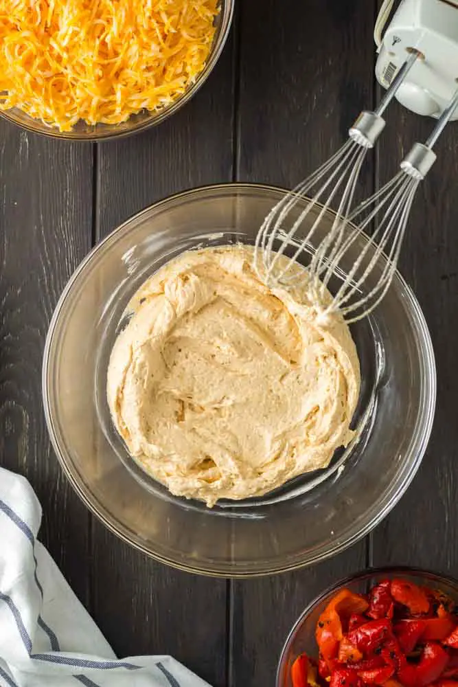 clear glass bowl filled with blended cream cheese, boursin cheese, mayonnaise, blackening seasoning and sriracha with bowls of shredded cheese and roasted red bell peppers around it