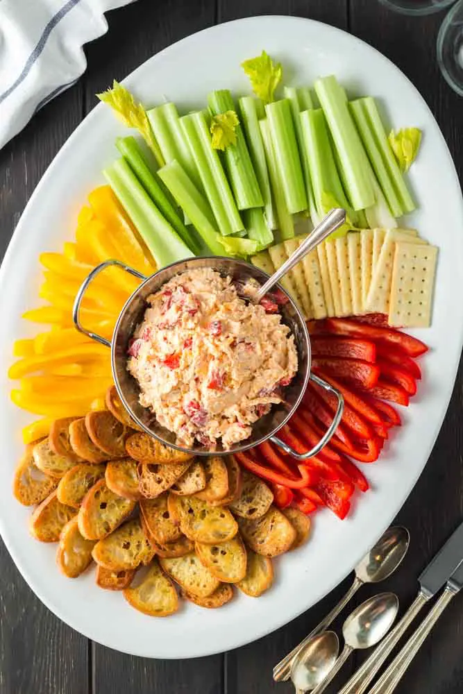 white oval shaped platter with a silver bowl of orange pimento cheese in the center surrounded by green celery sticks, red and yellow bell pepper strips and golden brown toasted baguette