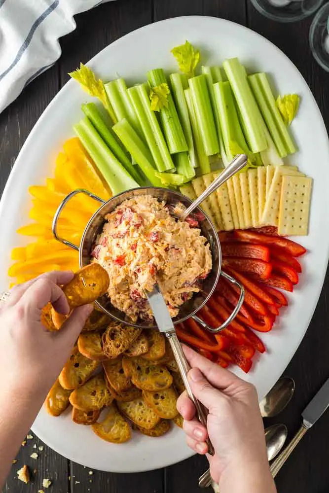 white oval shaped platter with a silver bowl of orange pimento cheese in the center surrounded by green celery sticks, red and yellow bell pepper strips and golden brown toasted baguette and hands reaching in to spread cheese on a toast point