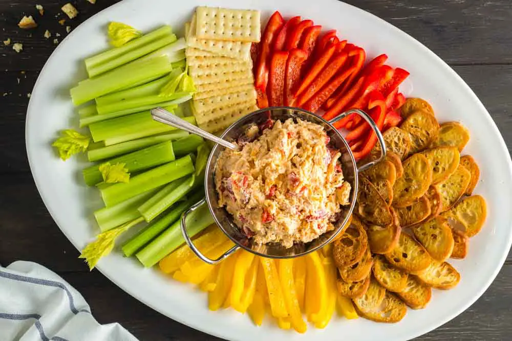 white oval shaped platter with a silver bowl of orange pimento cheese in the center surrounded by green celery sticks, red and yellow bell pepper strips and golden brown toasted baguette