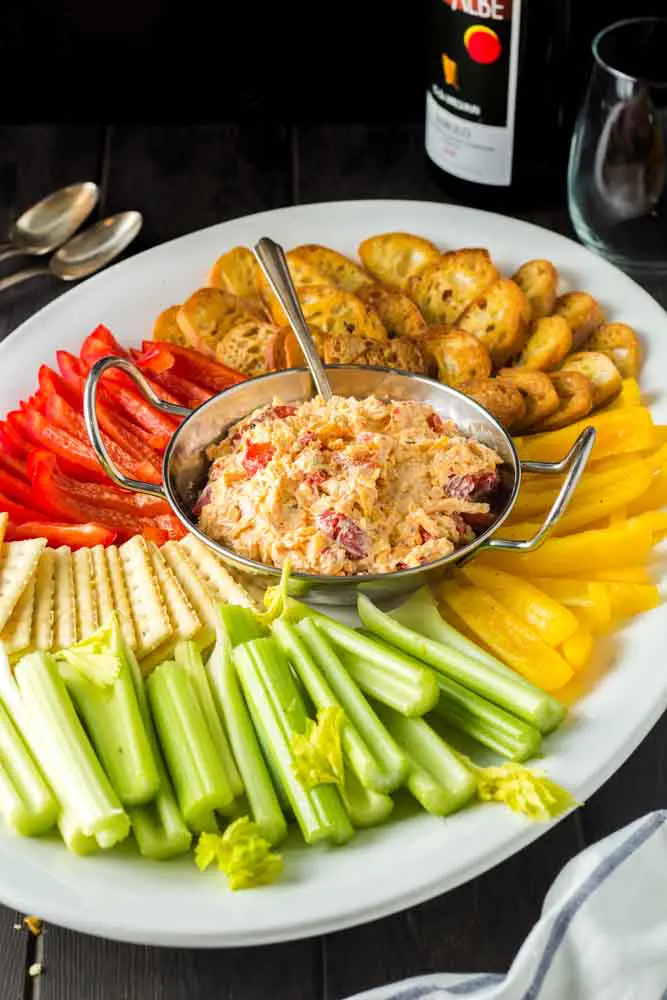 white oval shaped platter with a silver bowl of orange pimento cheese in the center surrounded by green celery sticks, red and yellow bell pepper strips and golden brown toasted baguette