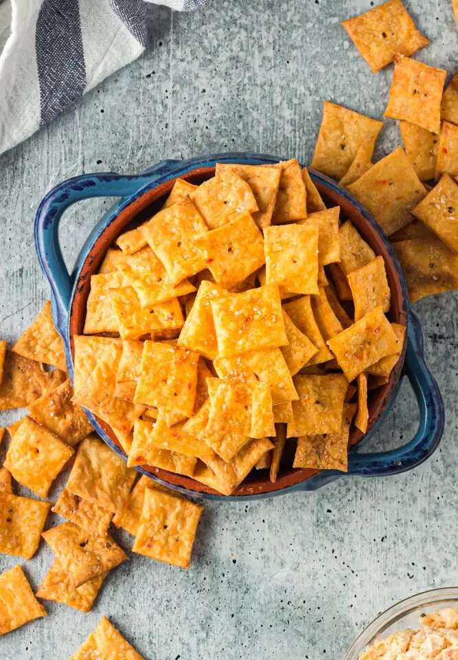 Bright orange square crackers with a single hole in the middle spilling out of a bright blue bowl