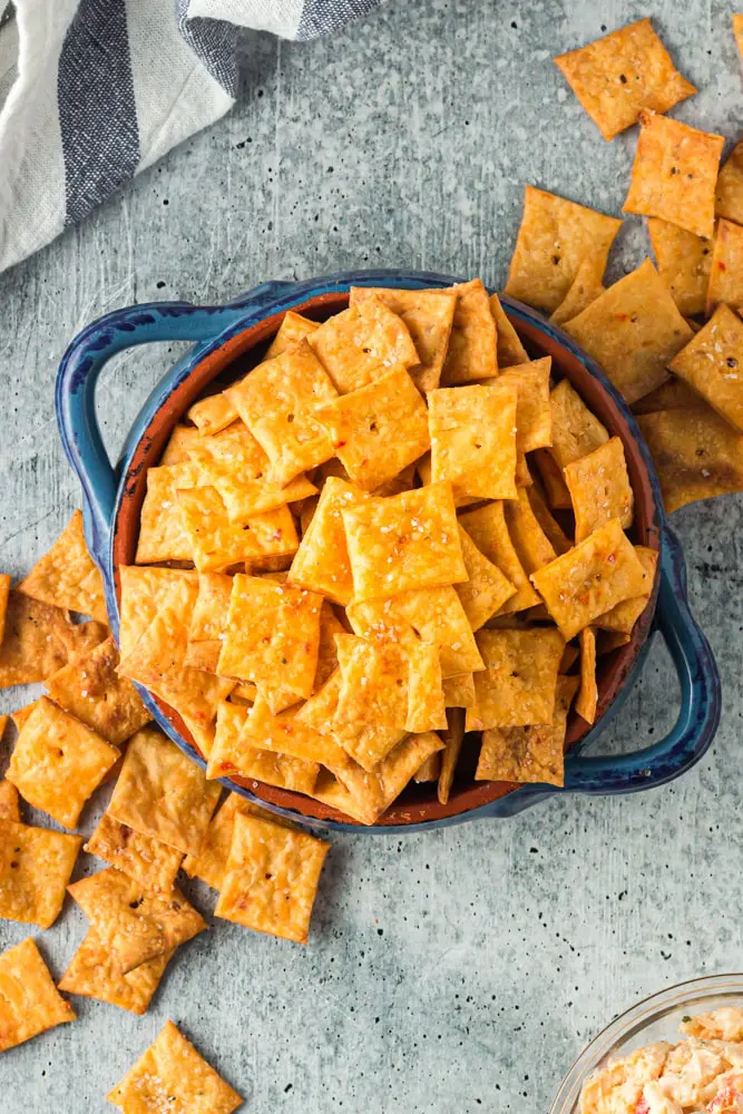 Bright orange square crackers with a single hole in the middle spilling out of a bright blue bowl