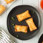 Golden brown rectangular pimento cheese wontons on small black plate and a cooling rack with a side of bright red thai sweet chili dipping sauce