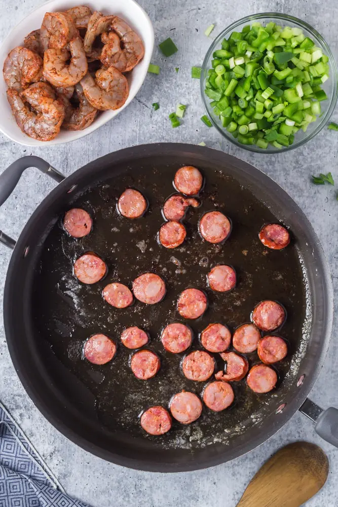 Large black saute pan willed with cooking andouille sausage rounds with a bowl of marinated shrimp and sliced green onions on the side