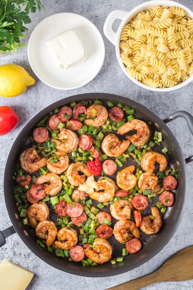 Sauteed shrimp and andouille sauage with green onions surrounded by small bowl of cream cheese and a strainer of cooked pasta