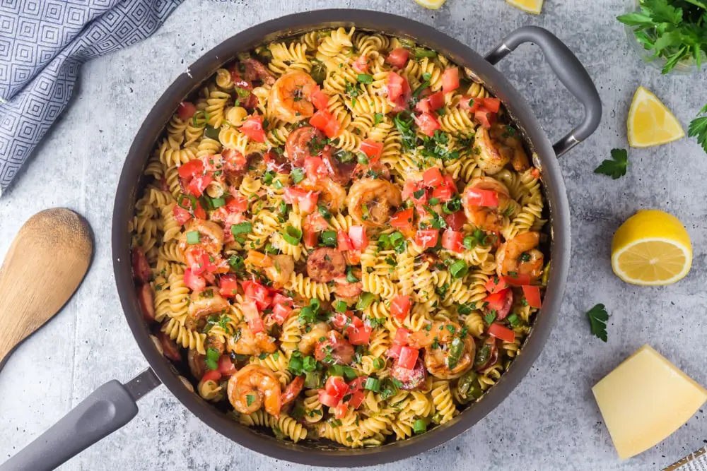 Large black saute pan filled with Cajun Shrimp Pasta with rotini pasta, andouille sausage rounds, bright red tomatoes and bell peppers and sliced green onions