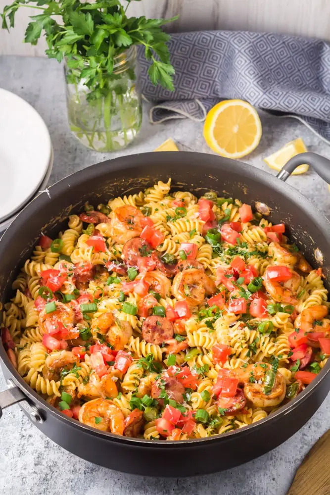 Large black saute pan filled with Cajun Shrimp Pasta with rotini pasta, andouille sausage rounds, bright red tomatoes and bell peppers and sliced green onions