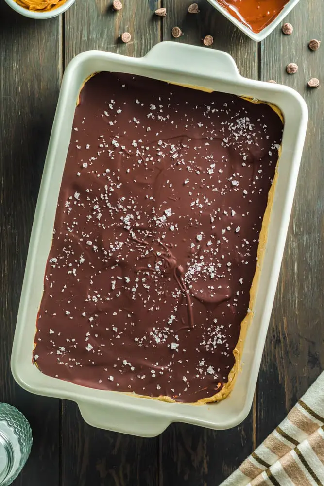 Chocolate Peanut Butter Bars made up of a base layer of peanut butter cookie, topped with smooth peanut butter filling and topped with a shiny chocolate ganache and flakes of sea salt being