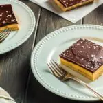 Chocolate Peanut Butter Bars made up of a base layer of peanut butter cookie, topped with smooth peanut butter filling and topped with a shiny chocolate ganache and flakes of sea salt being plated onto two white small plates