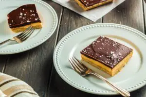 Chocolate Peanut Butter Bars made up of a base layer of peanut butter cookie, topped with smooth peanut butter filling and topped with a shiny chocolate ganache and flakes of sea salt being plated onto two white small plates