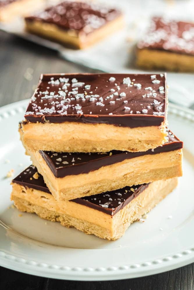 Three Chocolate Peanut Butter Bar squares on a small white plate- the squares are made up of a base layer of peanut butter cookie, topped with smooth peanut butter filling and topped with a shiny chocolate ganache and flakes of sea salt