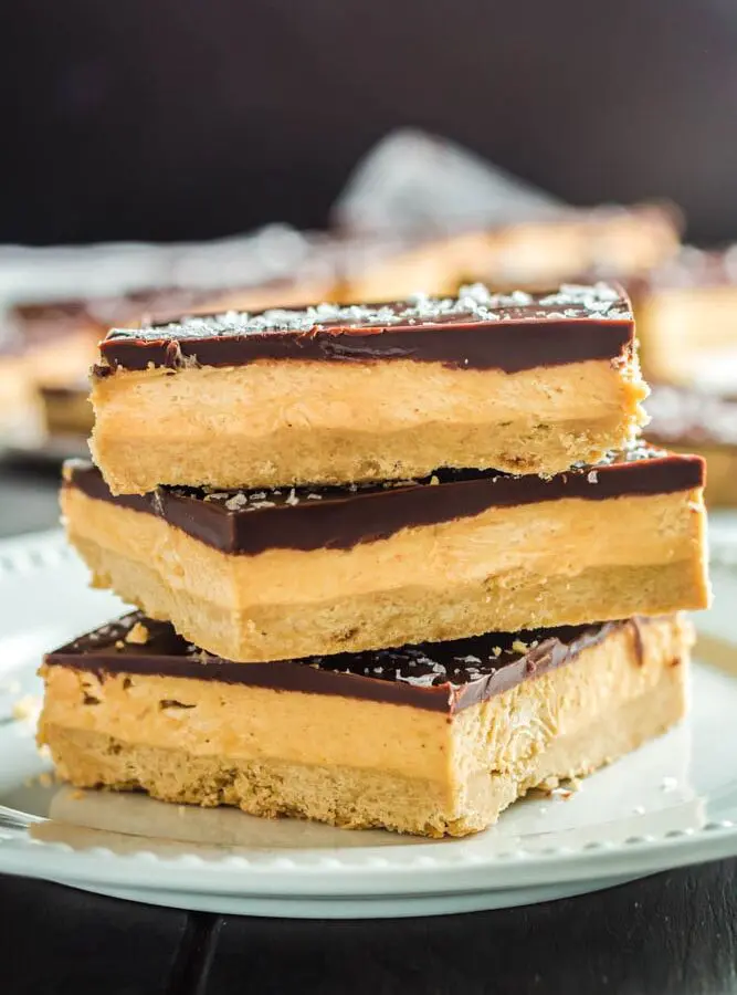 Three Chocolate Peanut Butter Bar squares on a small white plate- the squares are made up of a base layer of peanut butter cookie, topped with smooth peanut butter filling and topped with a shiny chocolate ganache and flakes of sea salt