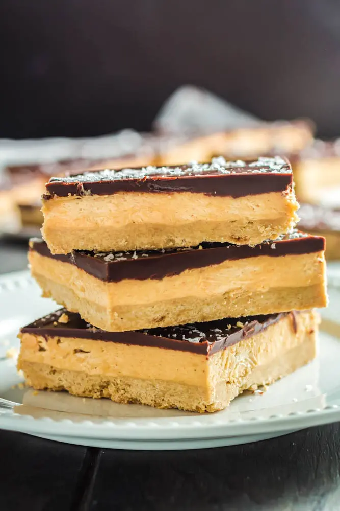 Three Chocolate Peanut Butter Bar squares on a small white plate- the squares are made up of a base layer of peanut butter cookie, topped with smooth peanut butter filling and topped with a shiny chocolate ganache and flakes of sea salt