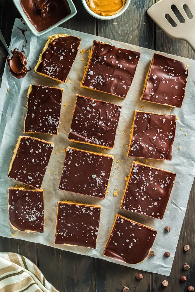 Chocolate Peanut Butter Bars made up of a base layer of peanut butter cookie, topped with smooth peanut butter filling and topped with a shiny chocolate ganache and flakes of sea salt cut into twelve squares on a piece of parchment paper