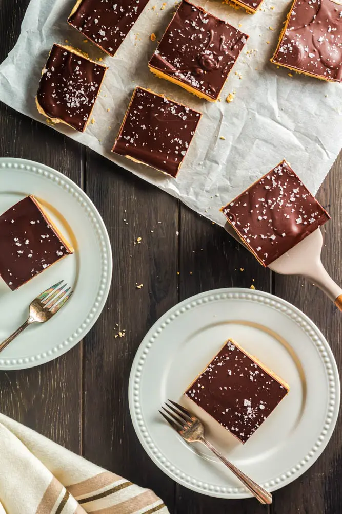 Chocolate Peanut Butter Bars made up of a base layer of peanut butter cookie, topped with smooth peanut butter filling and topped with a shiny chocolate ganache and flakes of sea salt being plated onto two white small plates