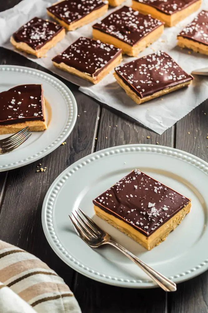 Chocolate Peanut Butter Bars made up of a base layer of peanut butter cookie, topped with smooth peanut butter filling and topped with a shiny chocolate ganache and flakes of sea salt being plated onto two white small plates