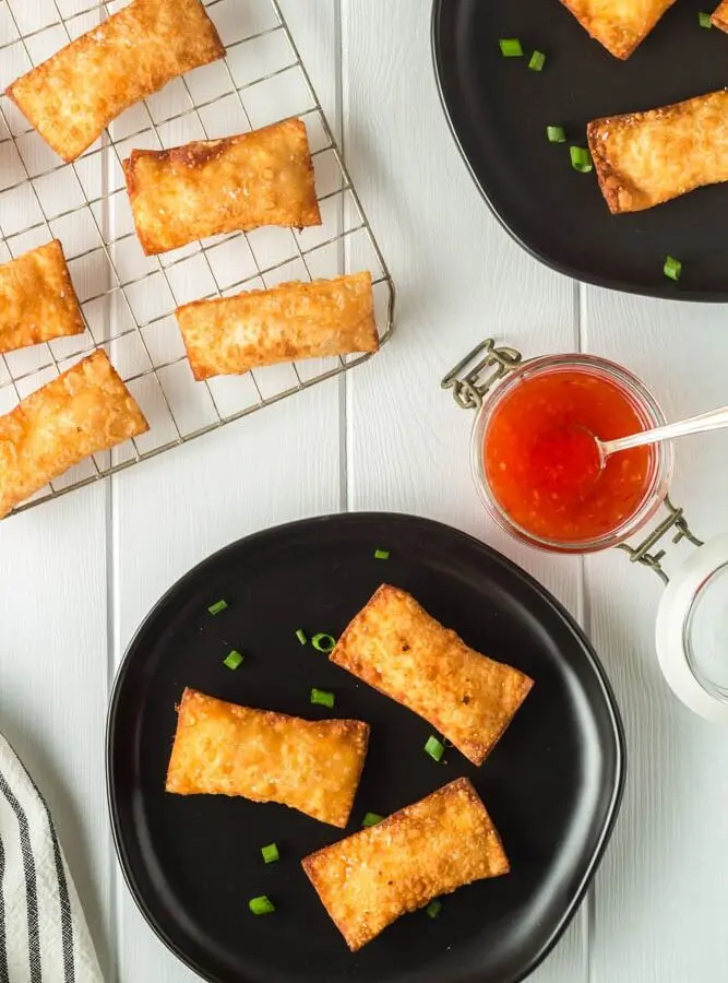 Golden brown rectangular pimento cheese wontons on small black plate and a cooling rack with a side of bright red thai sweet chili dipping sauce