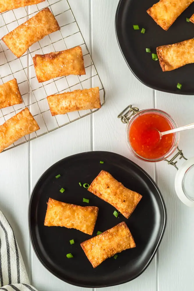 Golden brown rectangular pimento cheese wontons on small black plate and a cooling rack with a side of bright red thai sweet chili dipping sauce
